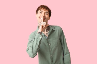 Mutlu, yakışıklı, dövmeli adam pembe arka planda sessiz jest yapıyor.