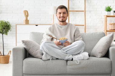 Yakışıklı genç adam elinde mısır gevreği yüzükleriyle oturma odasında kanepede oturuyor.