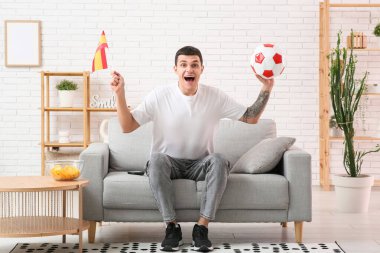 Futbol topu ve İspanya bayrağıyla evdeki kanepede futbol izleyen genç bir adam.