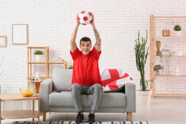 Bir kase patates cipsi, futbol topu, bir bardak bira ve evdeki kanepede futbol maçı izleyen İngiltere bayrağı olan genç adam.