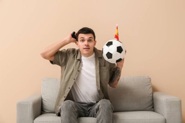 Futbol topu, televizyon kumandası ve bej duvarın yanındaki koltukta İspanya bayrağı olan genç bir adam.