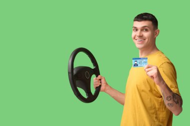 Yeşil arka planda ehliyet ve direksiyon tutan mutlu genç adam.