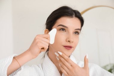 Bornozlu güzel bir kadın Guasha aracıyla yüz geliştirme egzersizi yapıyor.