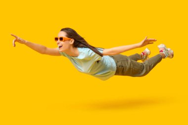 Güneş gözlüklü genç kadın uçuyor ve sarı arka plandaki bir şeyi işaret ediyor.