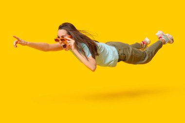 Güneş gözlüklü genç kadın uçuyor ve sarı arka plandaki bir şeyi işaret ediyor.