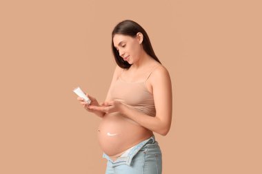 Genç hamile bir kadın bej arka planda çatlaklar için krem sürüyor.