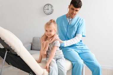 Erkek çocuk doktoru, klinikte steteskoplu küçük bir kızı muayene ediyor.