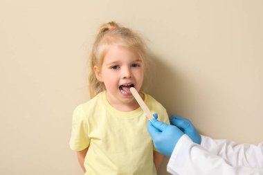 Pediatrician examining little girl with tongue depressor near beige wall