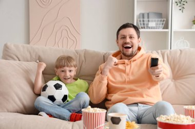 Genç baba ve küçük oğlu futbol topuyla evde maç izliyorlar.