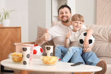 Genç baba ve küçük oğul evde maç izlerken başparmak kaldırıyorlar.