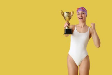 Altın kupalı mutlu genç bayan yüzücü sarı arka planda evet işareti yapıyor.