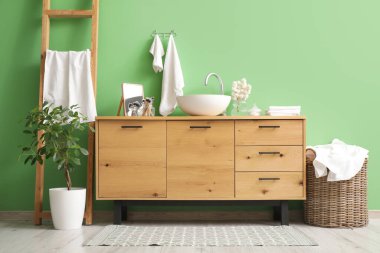 Modern banyodaki yeşil duvarın yanında lavabo ve çamaşır sepetli tahta sandık.