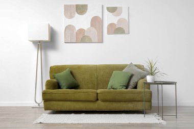 Modern oturma odasının içinde yeşil kanepe, lamba, sehpa ve beyaz duvarda resimler var.