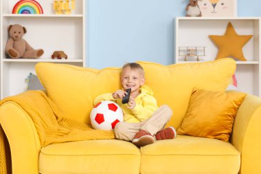 Sarı koltukta oturan, elinde futbol topu olan ve oyun odasında televizyon izleyen sevimli küçük çocuk.