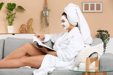 Evde yüz maskesi dergisi okuyan bornozlu genç bir Afro-Amerikalı kadın var.