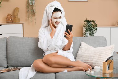 Yüz maskeli, bornozlu genç Afro-Amerikalı kadın evde selfie çekiyor.