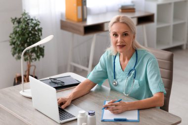 Klinikteki masada dizüstü bilgisayarla çalışan olgun bir kadın doktor.