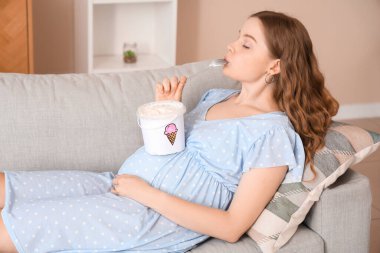 Evdeki kanepede dondurma yiyen hamile bir kadın.