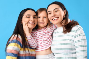 Mavi arka planda evlat edinilmiş küçük kızı kucaklayan genç lezbiyen çift.