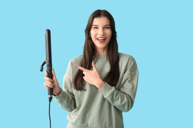 Güzel, genç ve mutlu bir kadın mavi arka planda saç maşasını işaret ediyor.