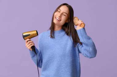 Saç kurutma makinesi ve leylak arkaplan fırçası olan mutlu genç bir kadın.