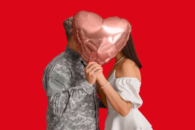 Askeri üniformalı bir adam ve karısı kırmızı arka planda kalp şeklinde bir balonun arkasında öpüşüyor. Sevgililer Günü kutlaması