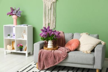 Raf ünitesinde bir buket leylak dalı, rahat bir kanepe ve ekoseli oturma odasının şık bir iç mekanı.