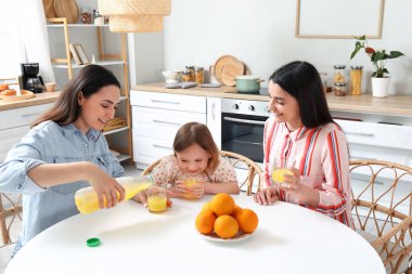 Evlatlık küçük kızı olan genç lezbiyen çift mutfakta portakal suyu içiyor.