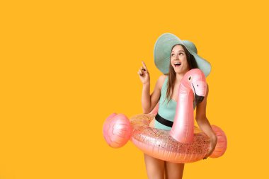 Havalı mayo giymiş, flamingo şeklinde şişirilebilir yüzüklü güzel bir kadın sarı arka plandaki bir şeyi işaret ediyor.