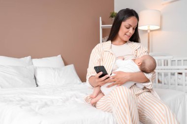Anne cep telefonuyla bebeğini yatak odasında emziriyor.