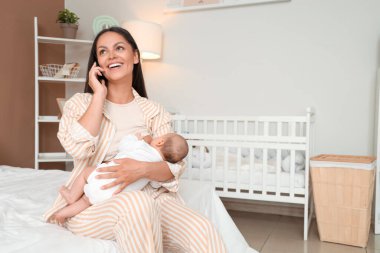 Anne yatak odasında cep telefonuyla konuşurken bebeğini emziriyor.