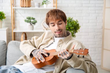 Havalı dövmeli genç adam evde ukulele çalıyor.