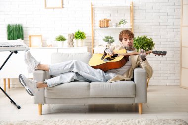 Evdeki kanepede otururken gitar çalan havalı dövmeli genç adam.