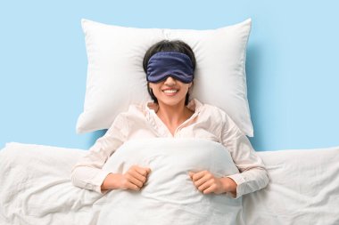 Mavi arka planda, üzerinde battaniye ve yumuşak yastık olan pijamalı güzel bir kadın.