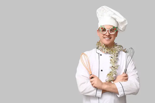 Stock image Portrait of young male chef with Christmas tinsel and whisks on grey background