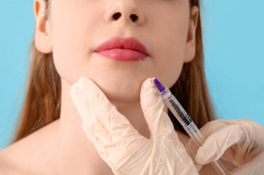 Genç bir kadın mavi arka planda yüzüne iğne vuruluyor.