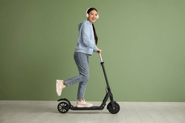 Kulaklıklı güzel bir Asyalı kadın yeşil duvarın yanında modern elektrikli scooter kullanıyor.