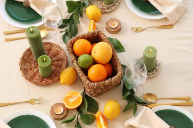 Narenciye meyveleri, yanan mumlar ve açık masa örtüsünün üzerindeki yeşil yapraklarla dolu güzel bir masa.