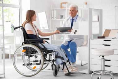 Kırık kollu tekerlekli sandalyedeki genç kadın hastanede doktoru ziyaret ediyor.