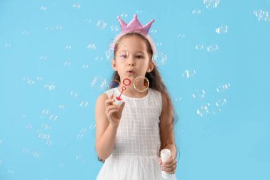 Mavi arka planda sabun köpüğü üfleyerek taç takan tatlı küçük kız.