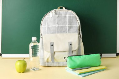 School backpack, stationery, bottle of water and apple on table against chalkboard