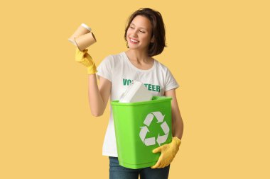 Sarı arka planda çöp ve kağıt bardaklarla geri dönüşüm kutusunu tutan kadın gönüllü.