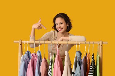 Güzel genç bir kadın sarı arka planda farklı giysiler olan rafları işaret ediyor.