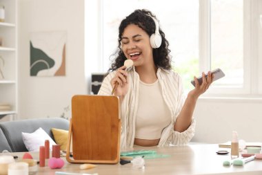 Makyaj fırçası ve cep telefonuyla evde şarkı söyleyen güzel, mutlu, Afro-Amerikan bir kadın.