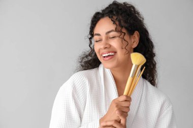 Gri arka planda makyaj fırçası olan bornozlu güzel, mutlu, Afro-Amerikan kadın.