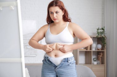 Şişman, üzgün bir kadın evinde aynadan karnına dokunuyor. Kilo alma kavramı