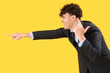 Gülen genç işadamı ezik jestler yapıyor ve sarı arka planda bir şeye işaret ediyor.