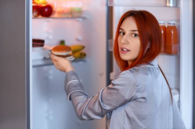 Genç bir kadın geceleri mutfaktaki buzdolabından lezzetli bir burger yiyor.