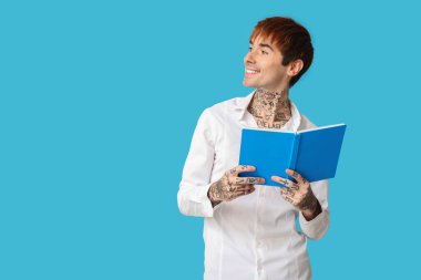 Mavi arka planda kitabı olan mutlu, yakışıklı, dövmeli bir adam.