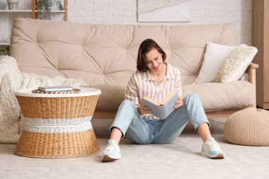 Genç, güzel bir kadın kitap okuyor ve oturma odasındaki kanepenin yanında oturuyor.